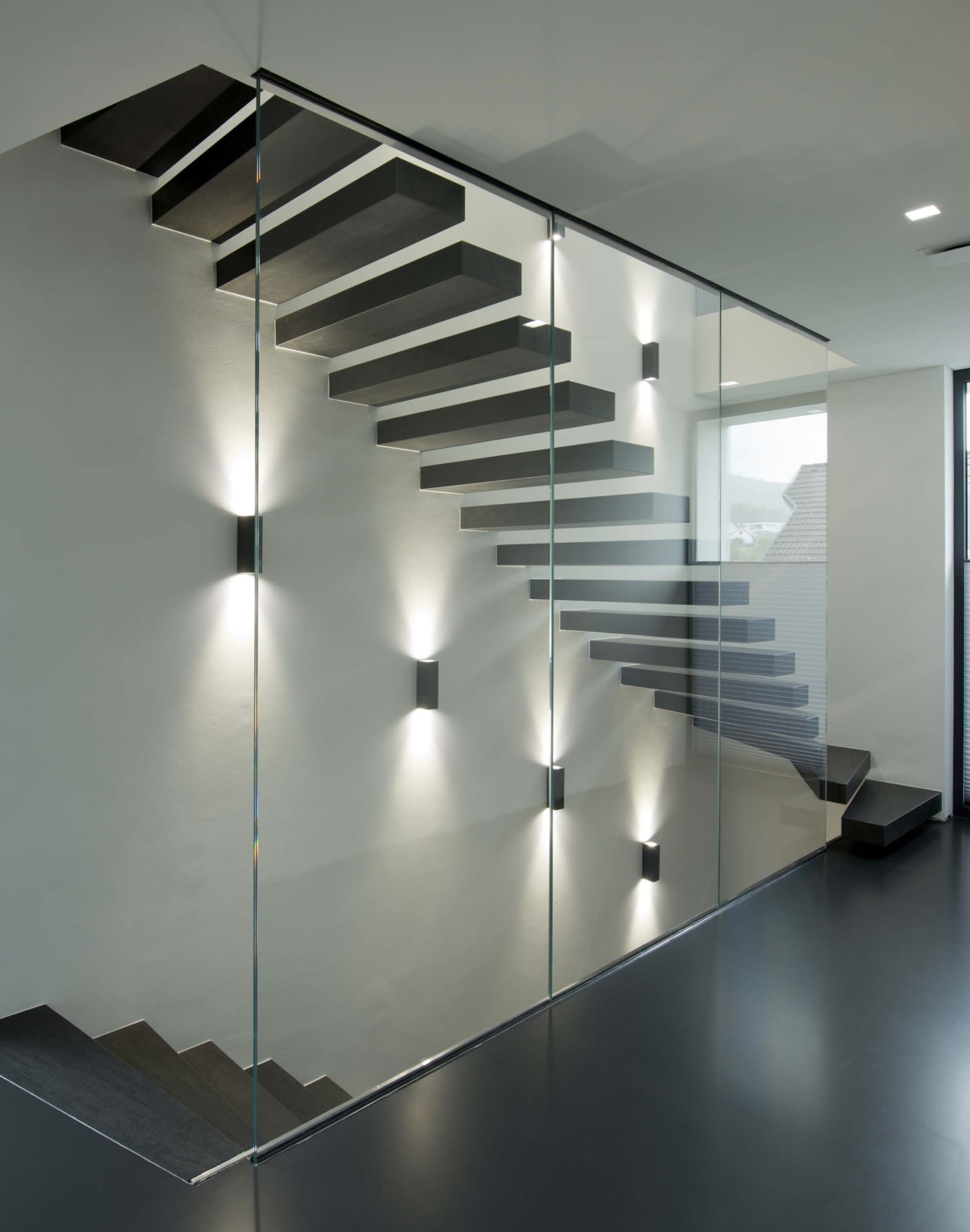 Kragarmtreppe mit dunklen Stufen hinter raumhoher Glaswand, Wandleuchten mit Licht nach oben und unten – AN Treppenbau