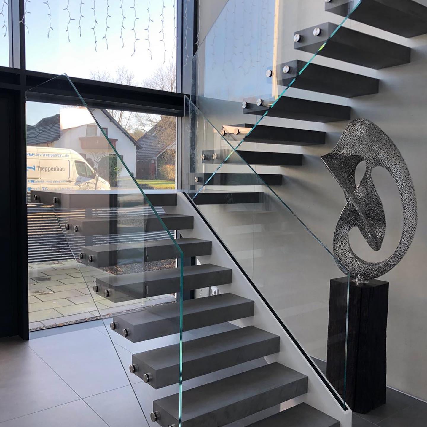 Kragarmtreppe mit grauen Stufen und rahmenlosem Glasgeländer vor einer hohen Fensterfront.