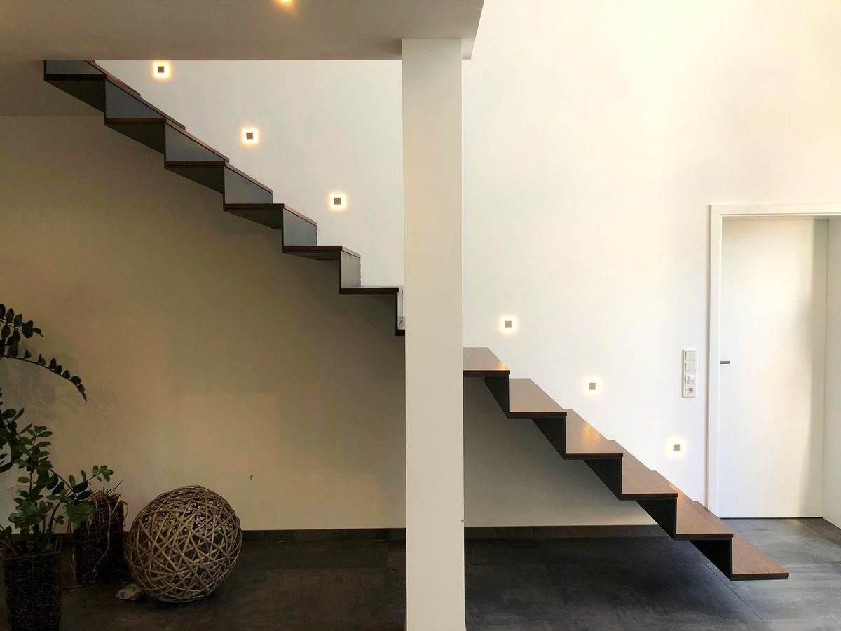 Faltwerktreppe aus Stahl mit Holzstufen an weißer Wand, entlang des Laufs quadratische Wandeinbauleuchten