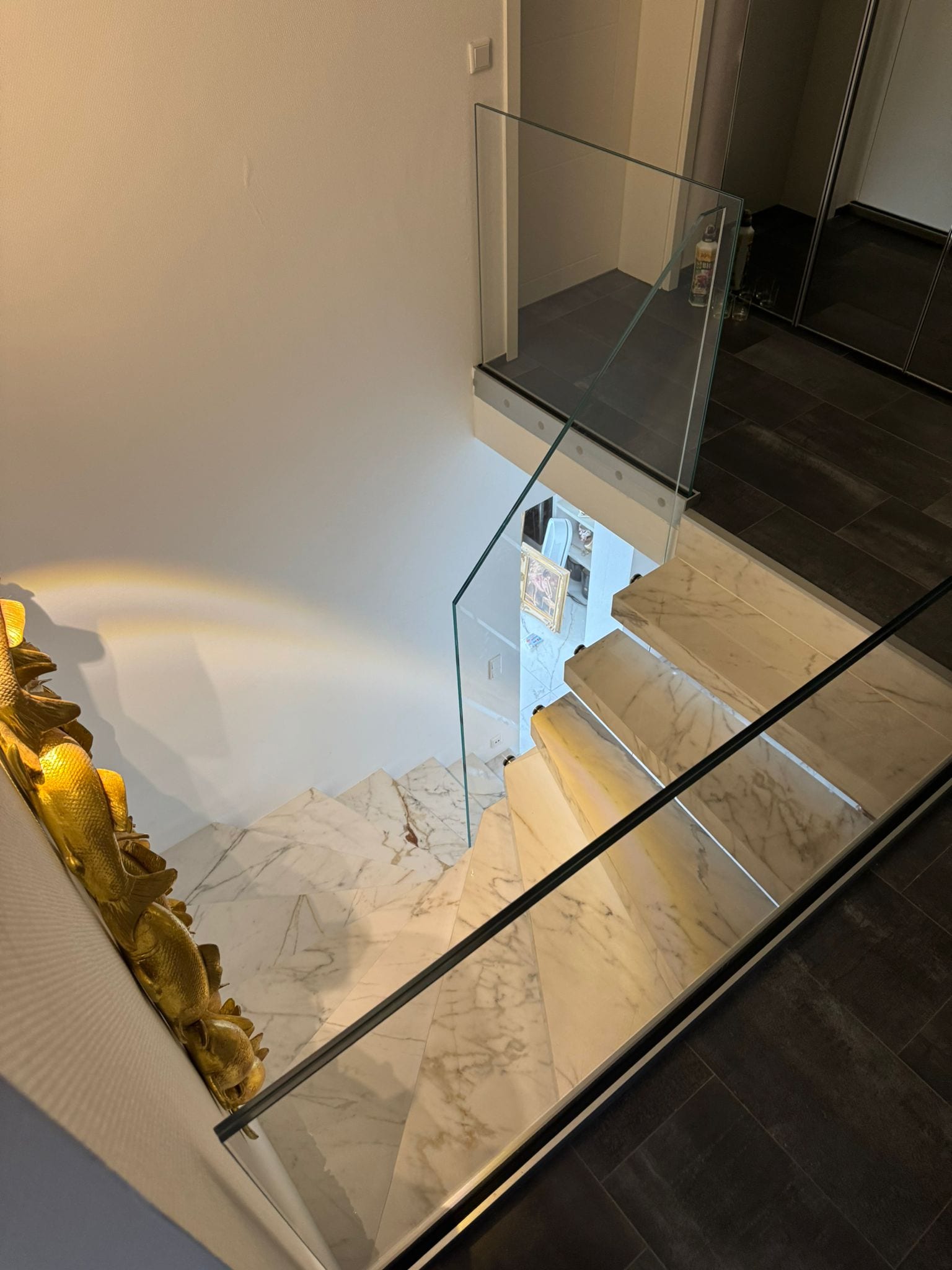 Blick von oben über das Ganzglasgeländer auf den unteren Treppenlauf mit Stufen in Marmoroptik – AN Treppenbau