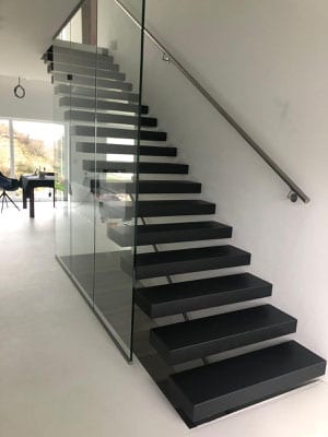 Kragarmtreppe mit schwarzen Keramikstufen, Ganzglasgeländer und Wandhandlauf im hellen Wohnraum – AN Treppenbau