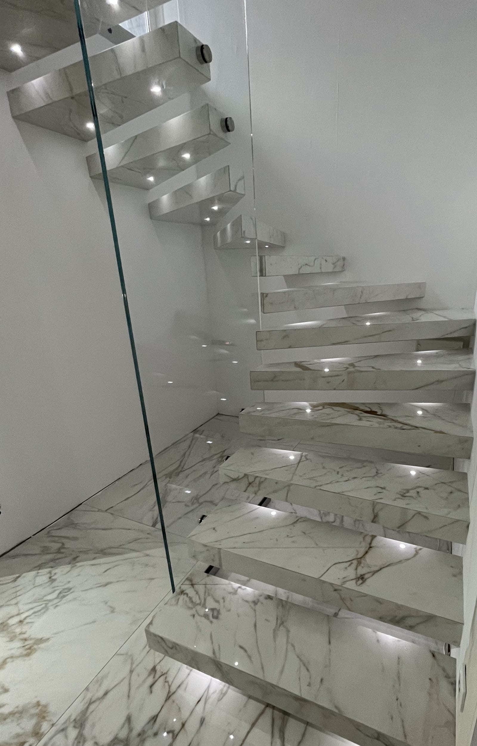 Kragarmtreppe mit hell gemaserten Stufen in Marmoroptik, Ganzglasgeländer und Punktleuchten – AN Treppenbau