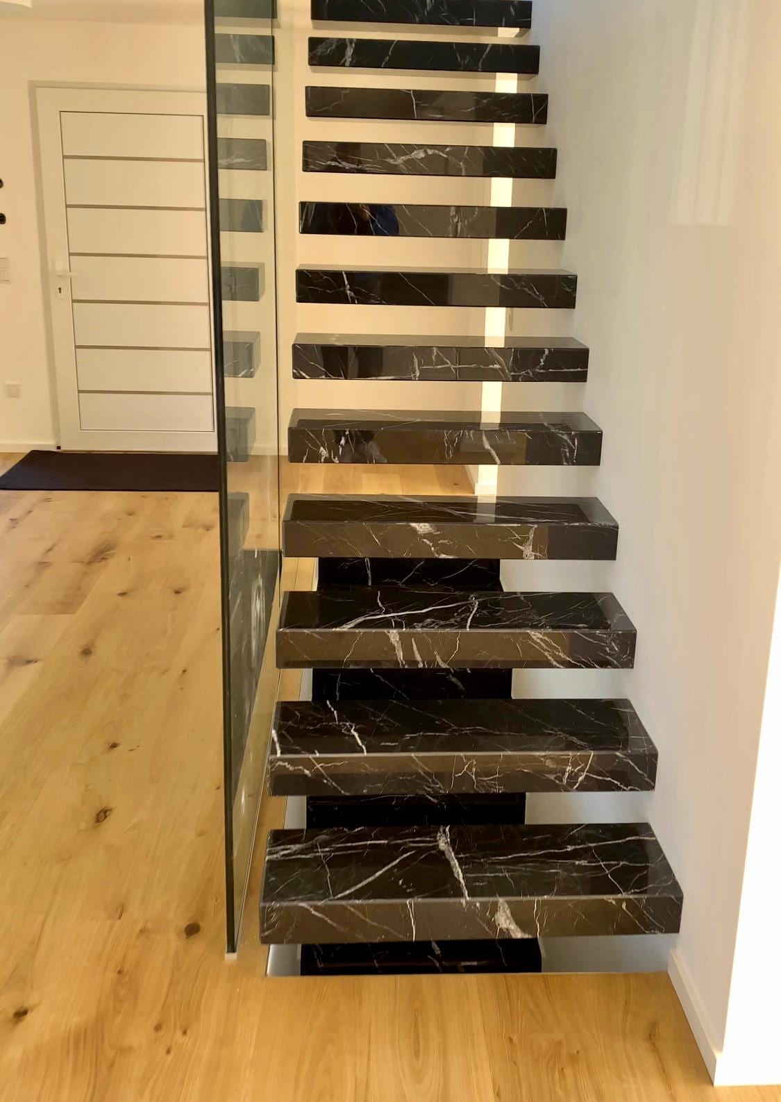 Kragarmtreppe mit schwarzen, weiß geaderten Steinstufen, Glasabtrennung und Lichtband an der Wand – AN Treppenbau