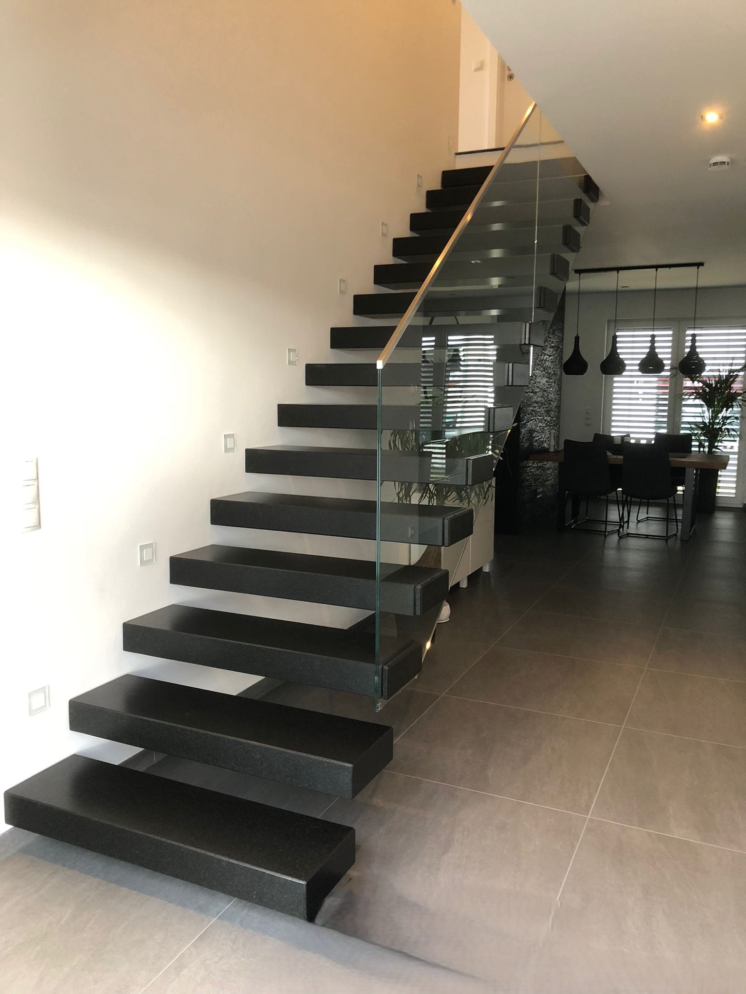 Kragarmtreppe mit anthrazitfarbenen Steinstufen, Glasgeländer mit hellem Handlauf, Blick zum Essbereich – AN Treppenbau
