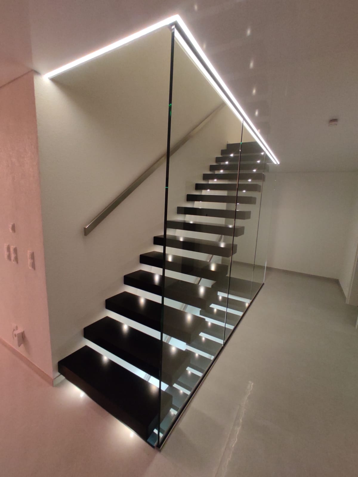 Dunkle Kragarmtreppe mit Punktleuchte in jeder Stufe, Ganzglasgeländer und Lichtband über dem Lauf – AN Treppenbau