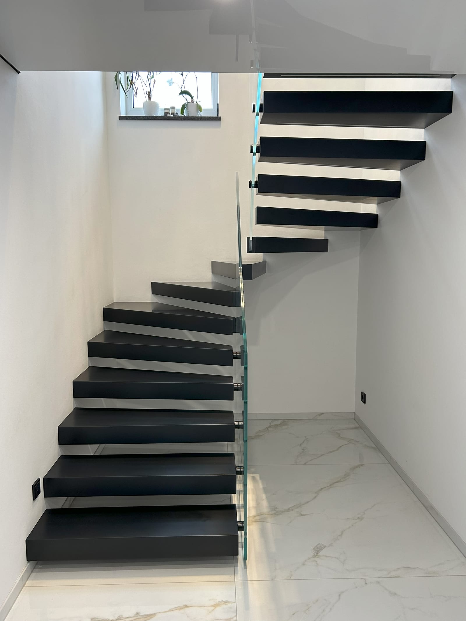 Gewendelte Kragarmtreppe mit dunklen Stufen und Ganzglasgeländer, Blick vom Treppenfuß über beide Läufe – AN Treppenbau