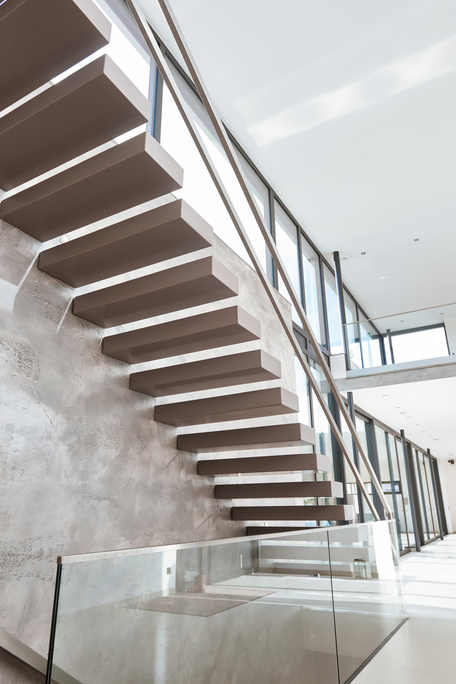 Kragarmtreppe mit hellgrauen Stufen und Glasgeländer vor einer Wand in Betonoptik, Blick von unten in ein Treppenhaus mit großen Fenstern