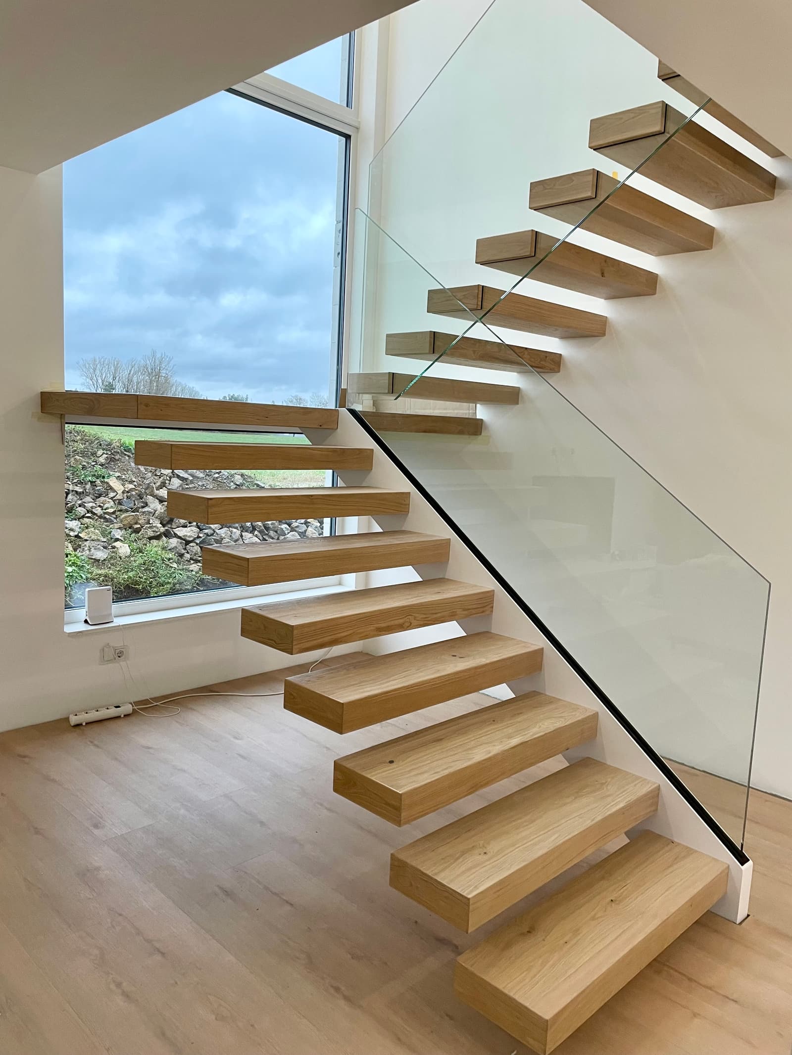 Kragarmtreppe aus Eiche mit rahmenlosem Glasgeländer vor einem großen Fenster, geölte Massivholzstufen