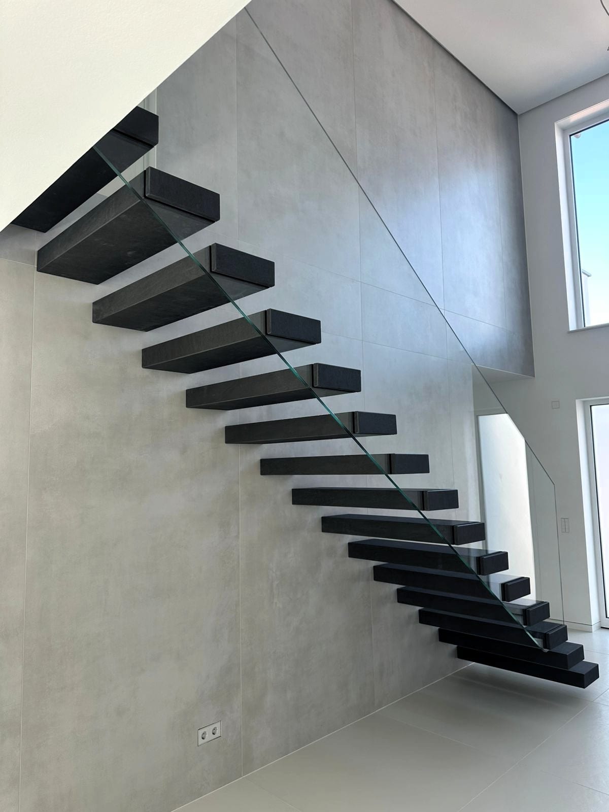 Kragarmtreppe mit anthrazitfarbenen Blockstufen und Ganzglasgeländer, Wand in Betonoptik – AN Treppenbau