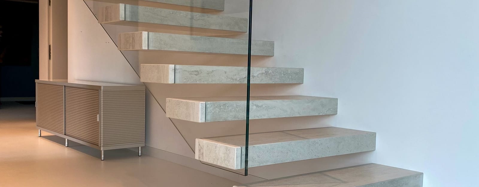 Kragarmtreppe mit hellen Stufen in Travertin-Optik und rahmenlosem Glasgeländer, Ansicht vom Fuß der Treppe