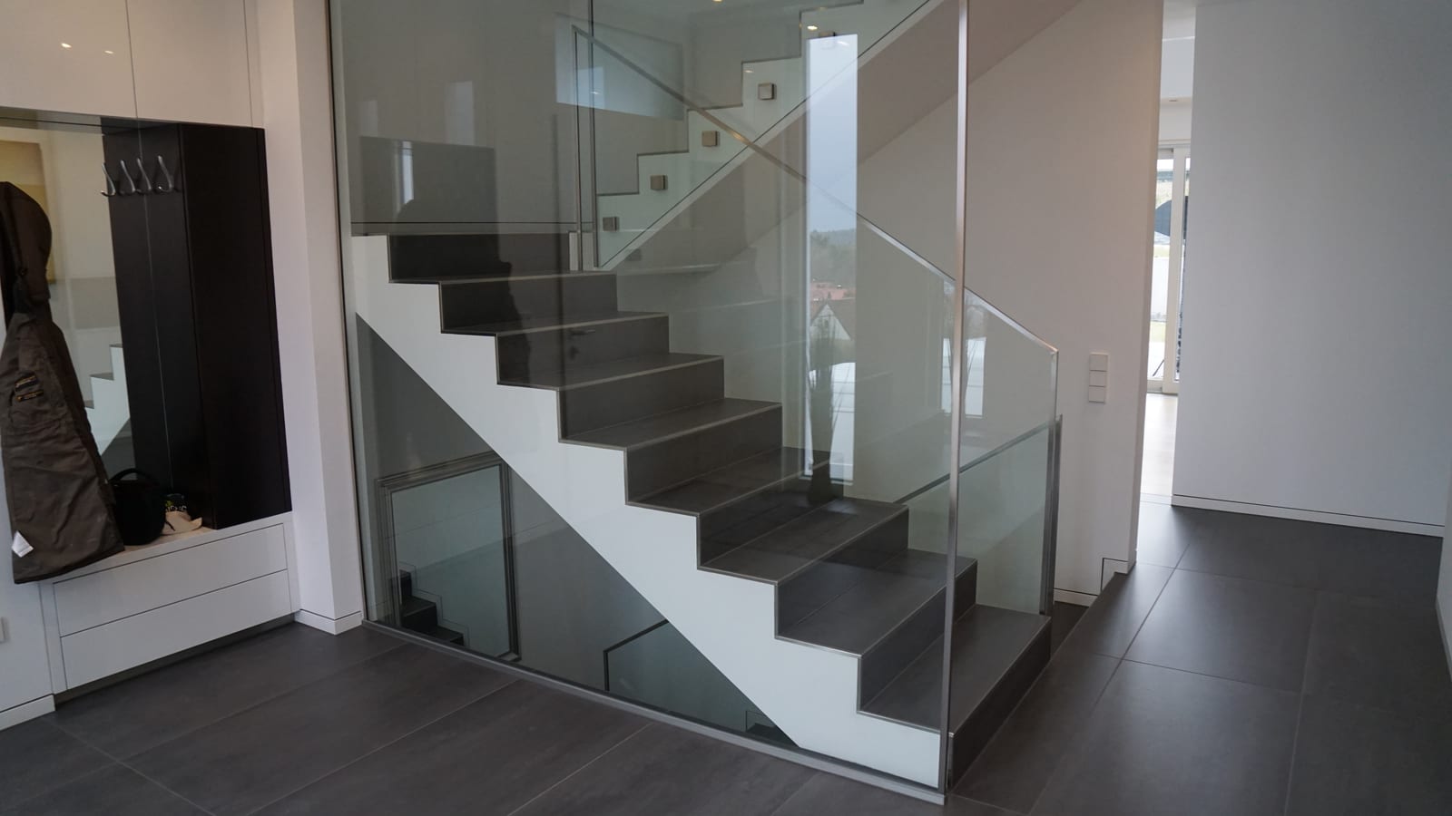 Kragarmtreppe mit dunklen Stufen und Ganzglasgeländer neben einer Garderobe