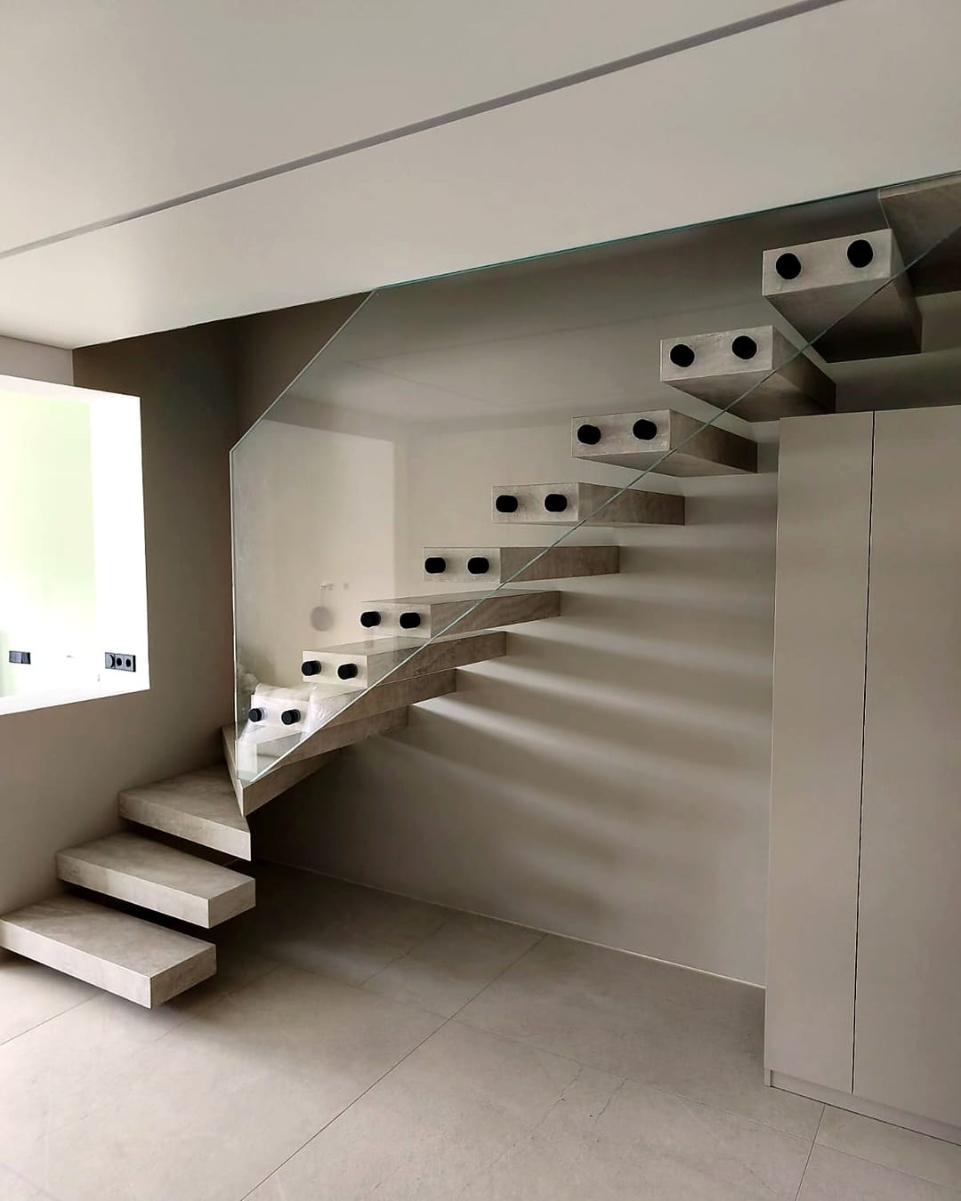 Freitragende Kragarmtreppe mit Stufen in heller Betonoptik und Ganzglasgeländer, gewendelter Antritt – AN Treppenbau
