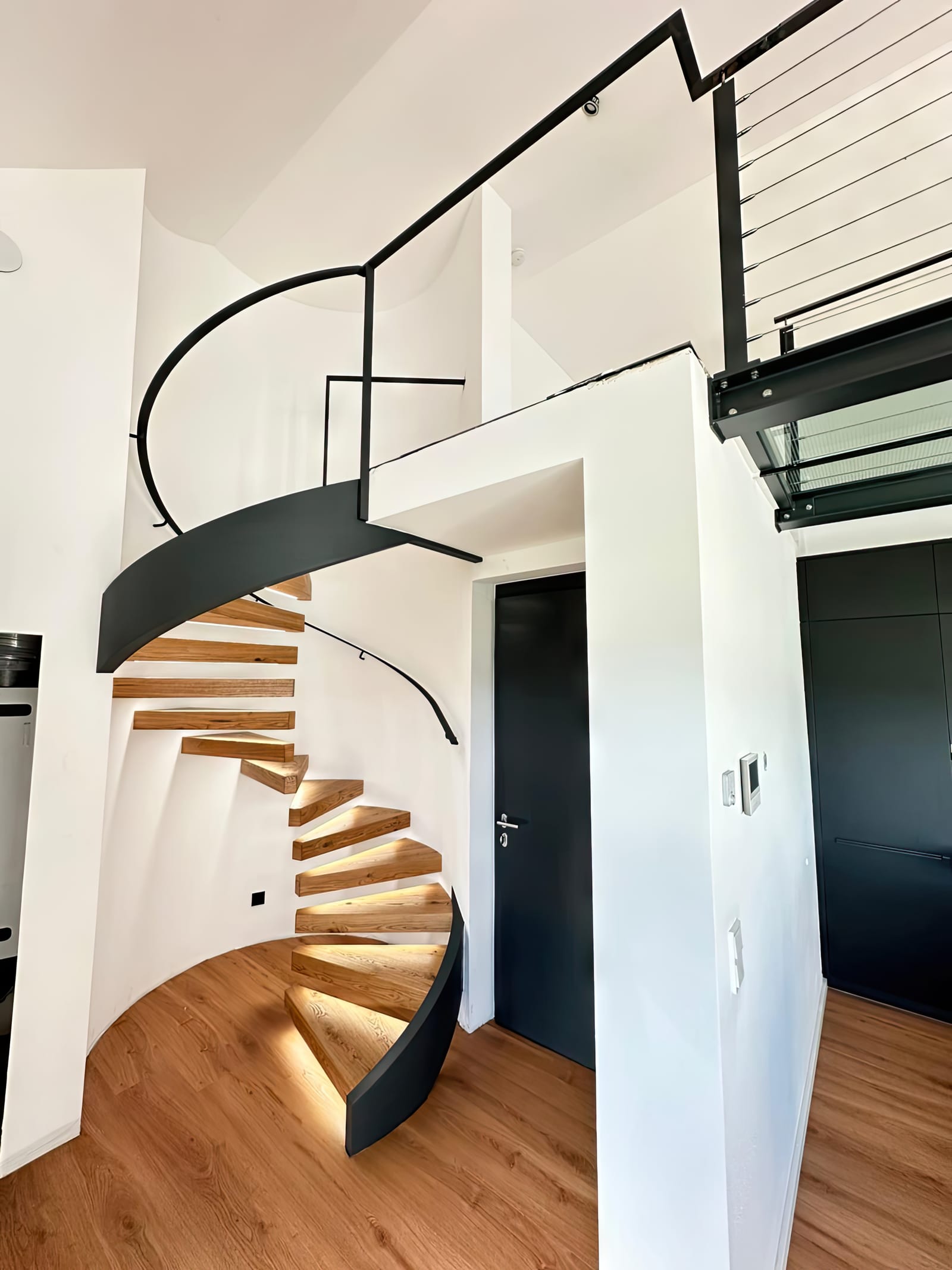 Gewendelte Kragarmtreppe mit beleuchteten Holzstufen und schwarzem Handlauf – AN Treppenbau